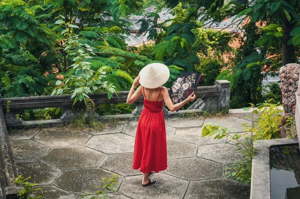 A woman in a long red dress and a straw hat walking through a leafy garden courtyard.
