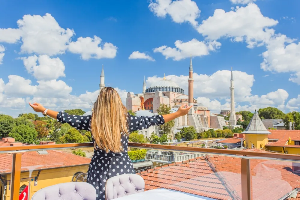 A woman in a polka-dot dress with her arms open on a rooftop terrace, looking out over the domes and minarets of Istanbul.