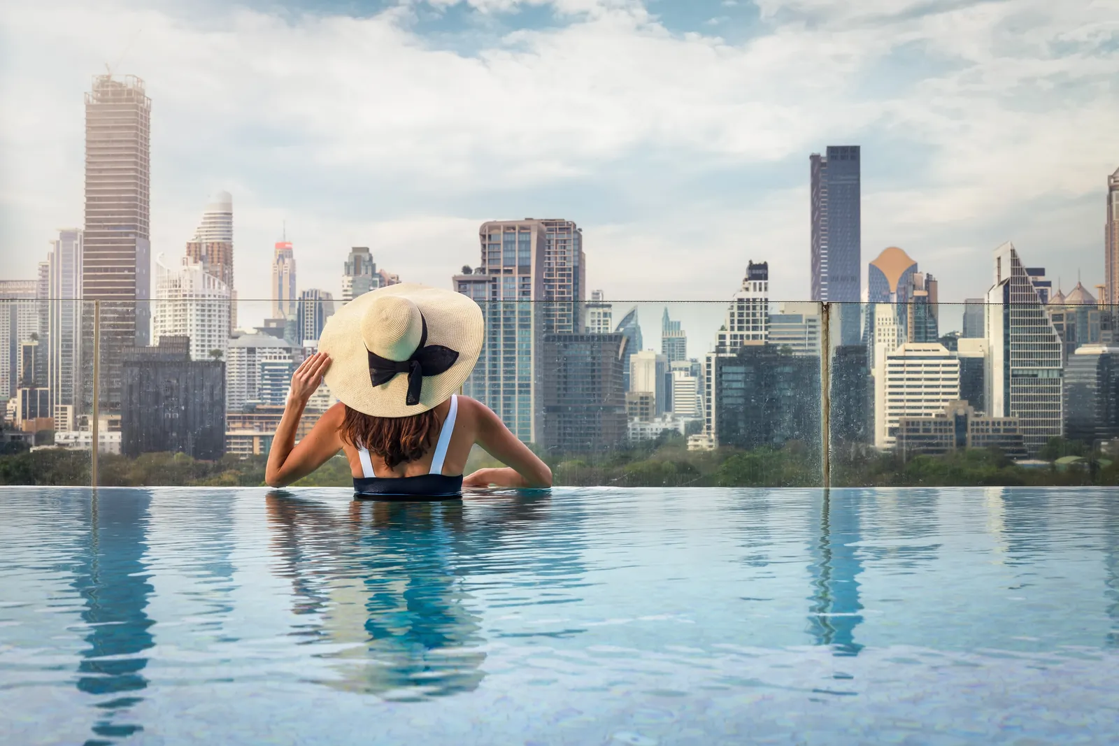 A woman in a sun hat in a rooftop pool, looking out over the Bangkok skyline