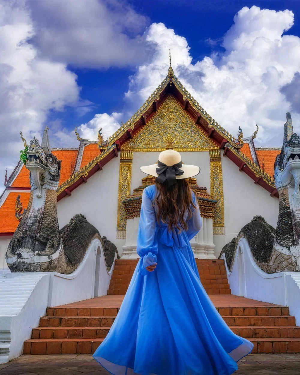 A woman in a blue dress walking up the steps of a Bangkok temple, golden spires ahead of her.